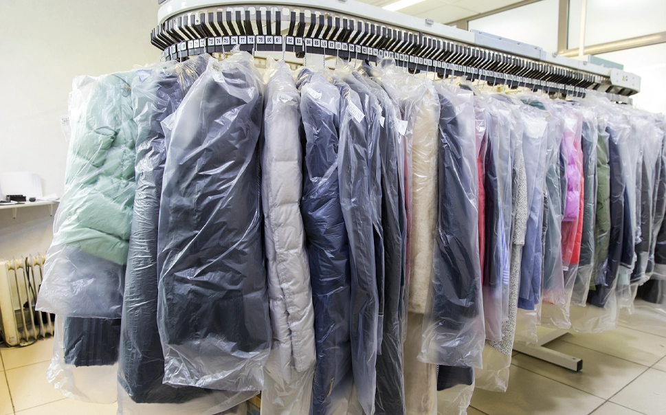 A row of cleaned clothes covered in plastic hanging on a conveyor rack in a dry cleaning facility.