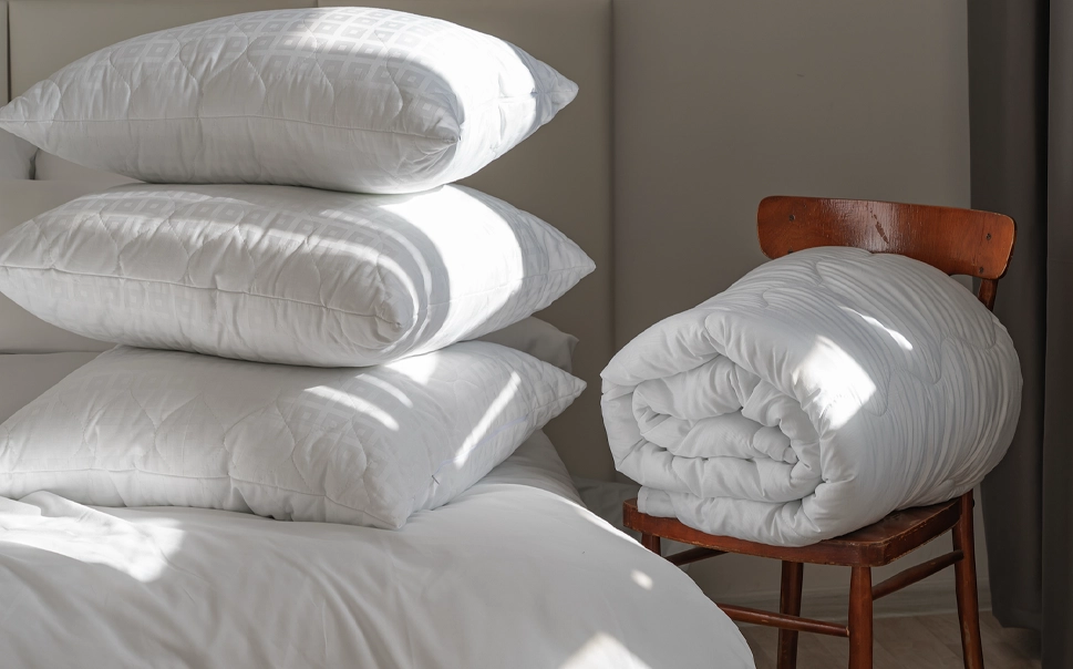 Three white pillows are stacked on a bed, while a rolled-up white comforter rests on a wooden chair beside the bed.
