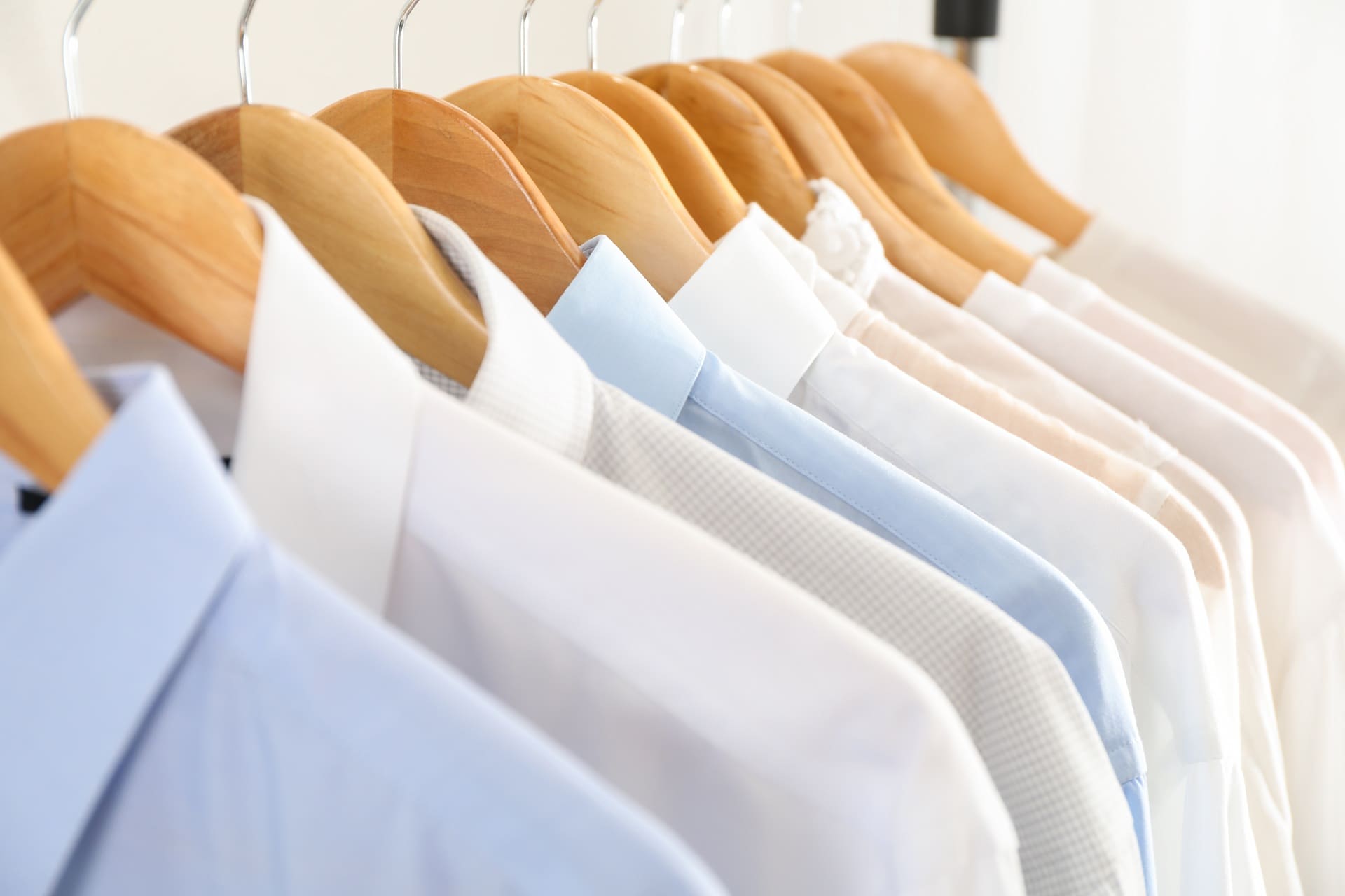 Several neatly pressed dress shirts in shades of white, blue, and beige hang on wooden hangers in a row.