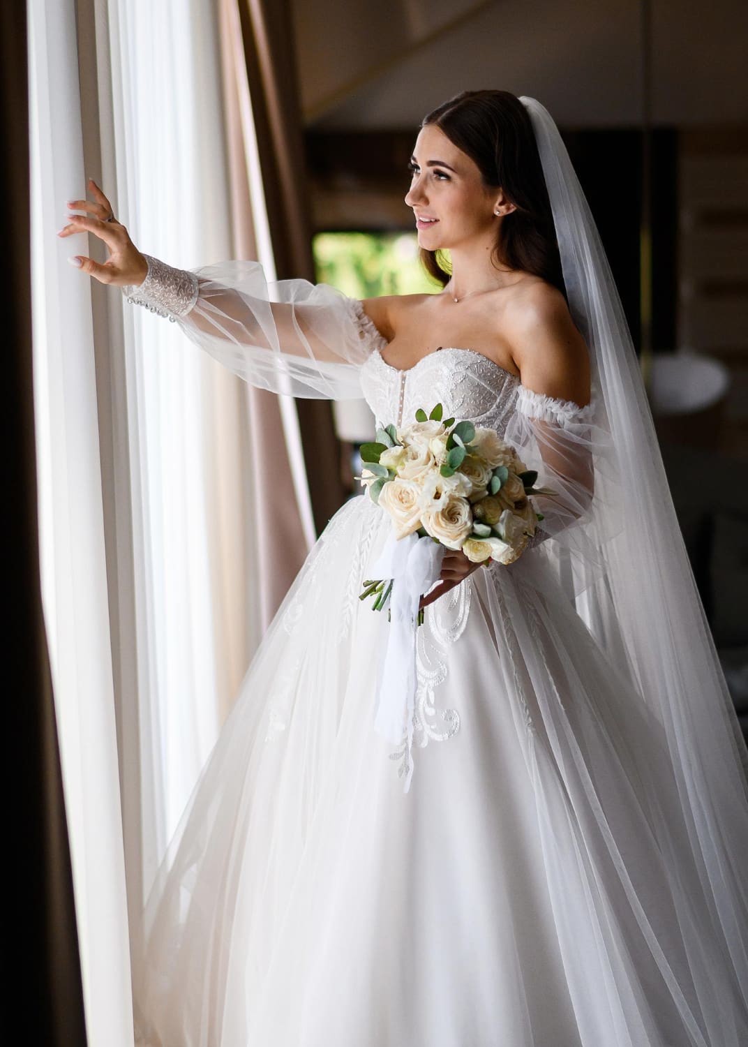 A bride in a white off-the-shoulder wedding dress and veil holds a bouquet of white roses while looking out a window.