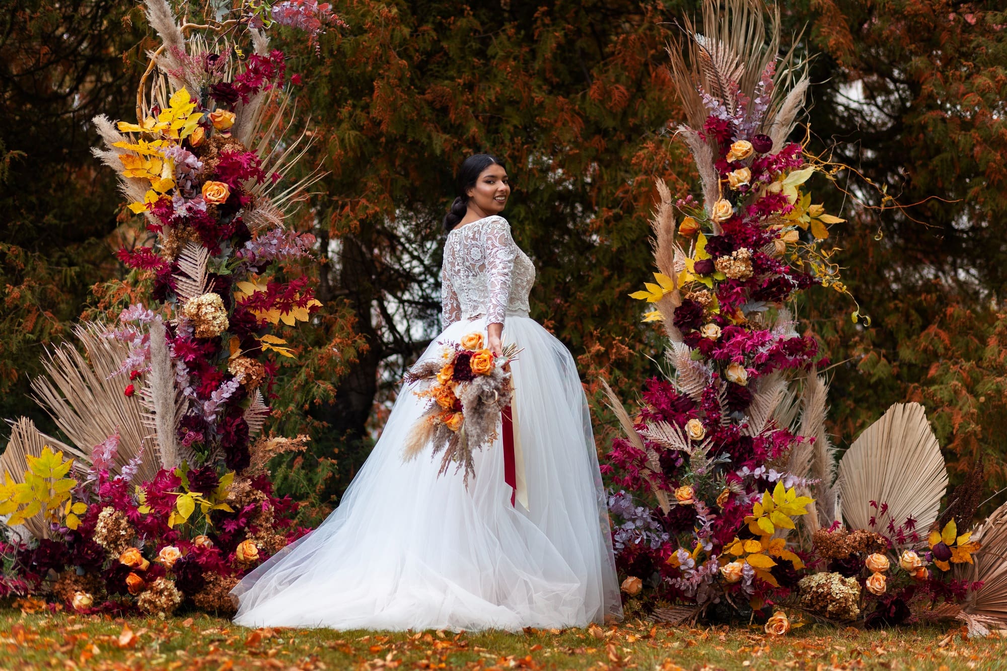A bride in a white wedding dress stands outdoors, holding a bouquet, surrounded by colorful autumn-themed floral arrangements.