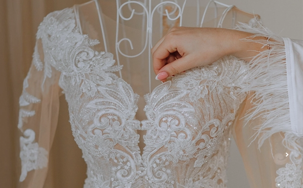 A hand adjusts the neckline of a white, intricately embroidered lace wedding dress displayed on a mannequin.