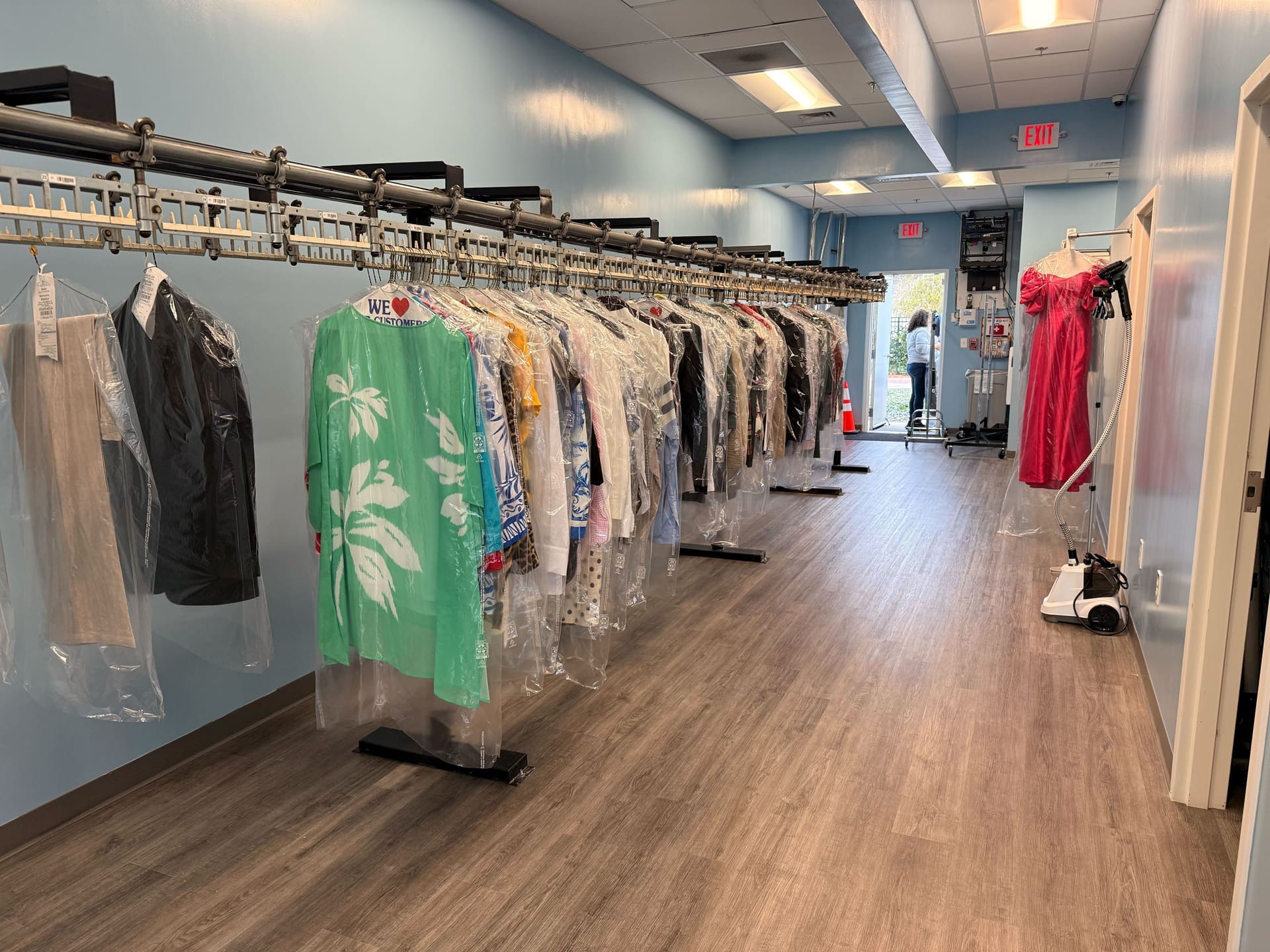 A dry cleaning shop interior with clothes on hangers covered in plastic along a conveyor rack, wood-like flooring, and a red dress on a mannequin near the entrance.