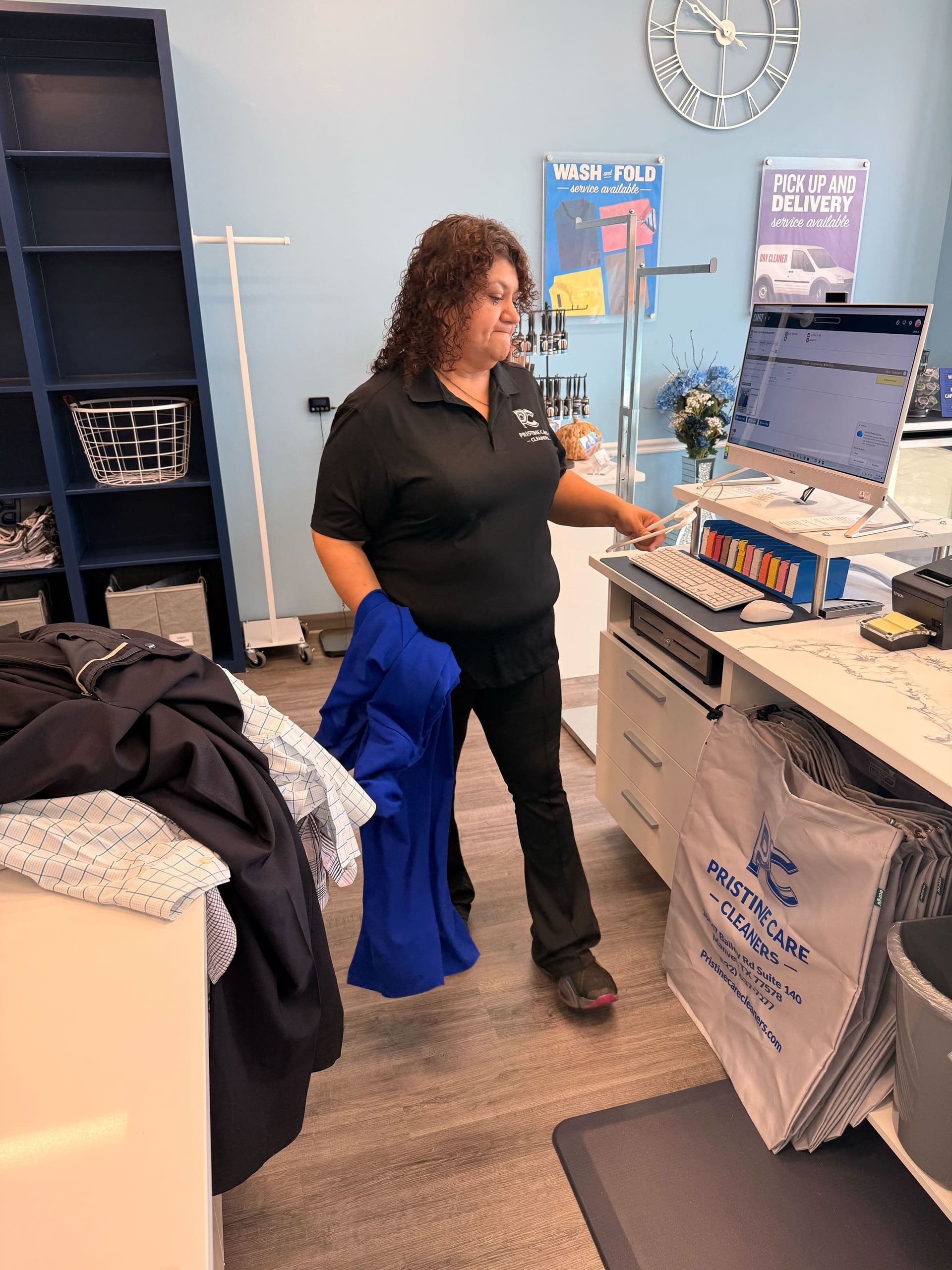 A woman in a black uniform stands at a dry cleaning counter, holding clothes and using a computer. Garment bags and folded laundry are visible around her.