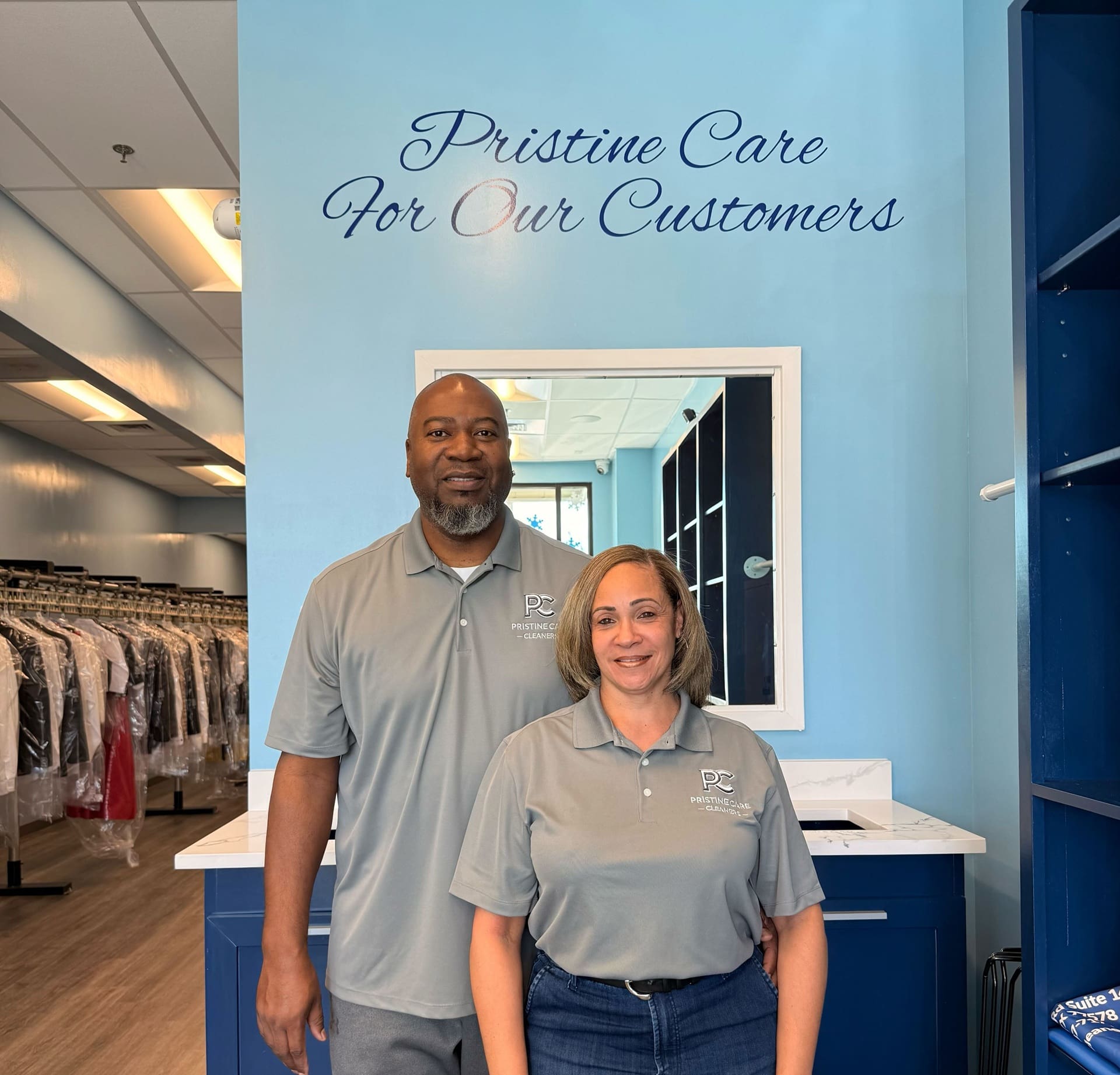 Two people in matching gray shirts stand inside a dry cleaning business with "Pristine Care For Our Customers" written on the wall behind them.