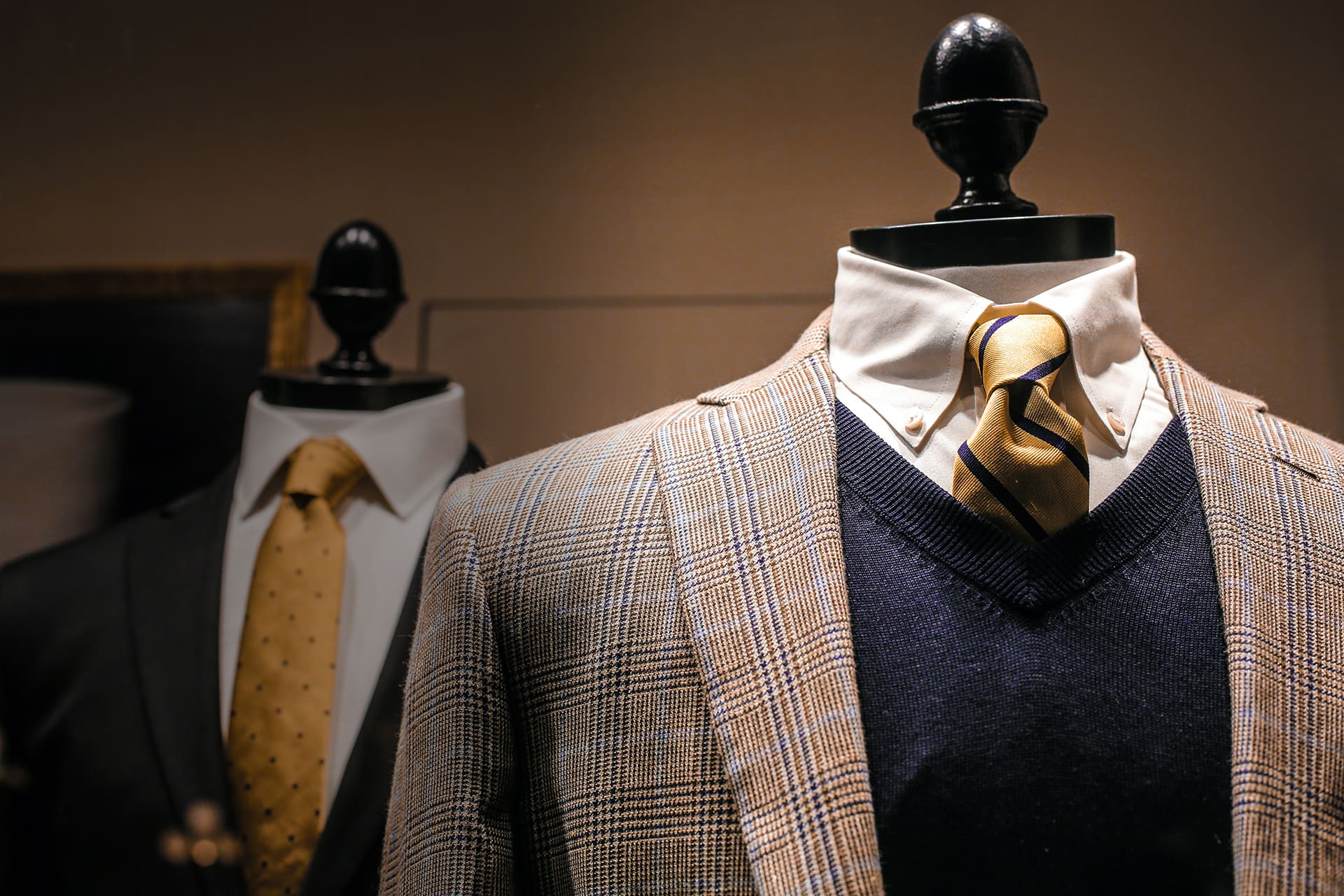 Two mannequins display men's suits; the front mannequin wears a checked blazer, navy sweater, white shirt, and gold tie, while the rear mannequin wears a dark suit and gold patterned tie.