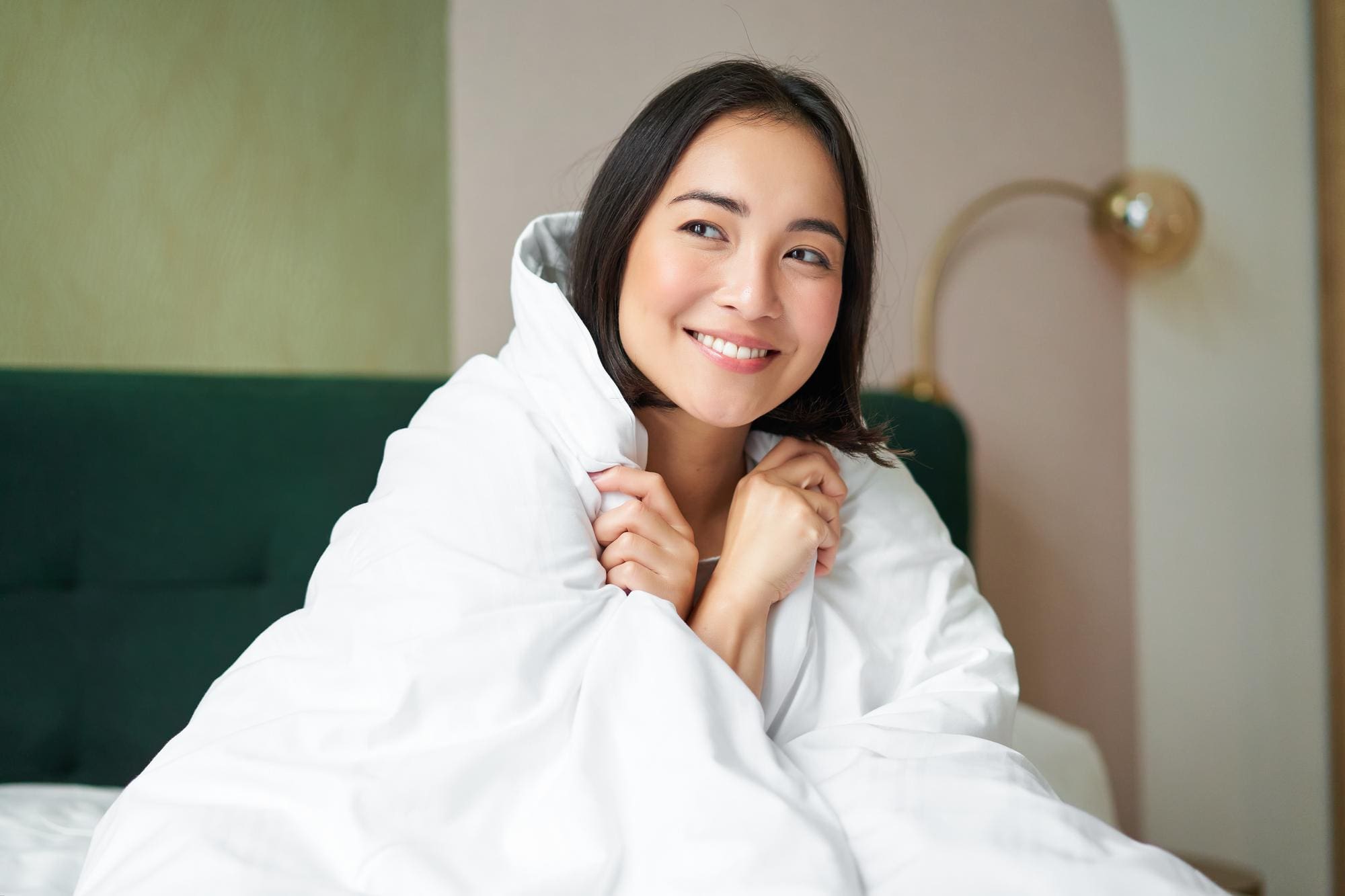 A woman sits on a bed, wrapped in a white blanket, smiling and looking to the side. A lamp and a green headboard are visible in the background.