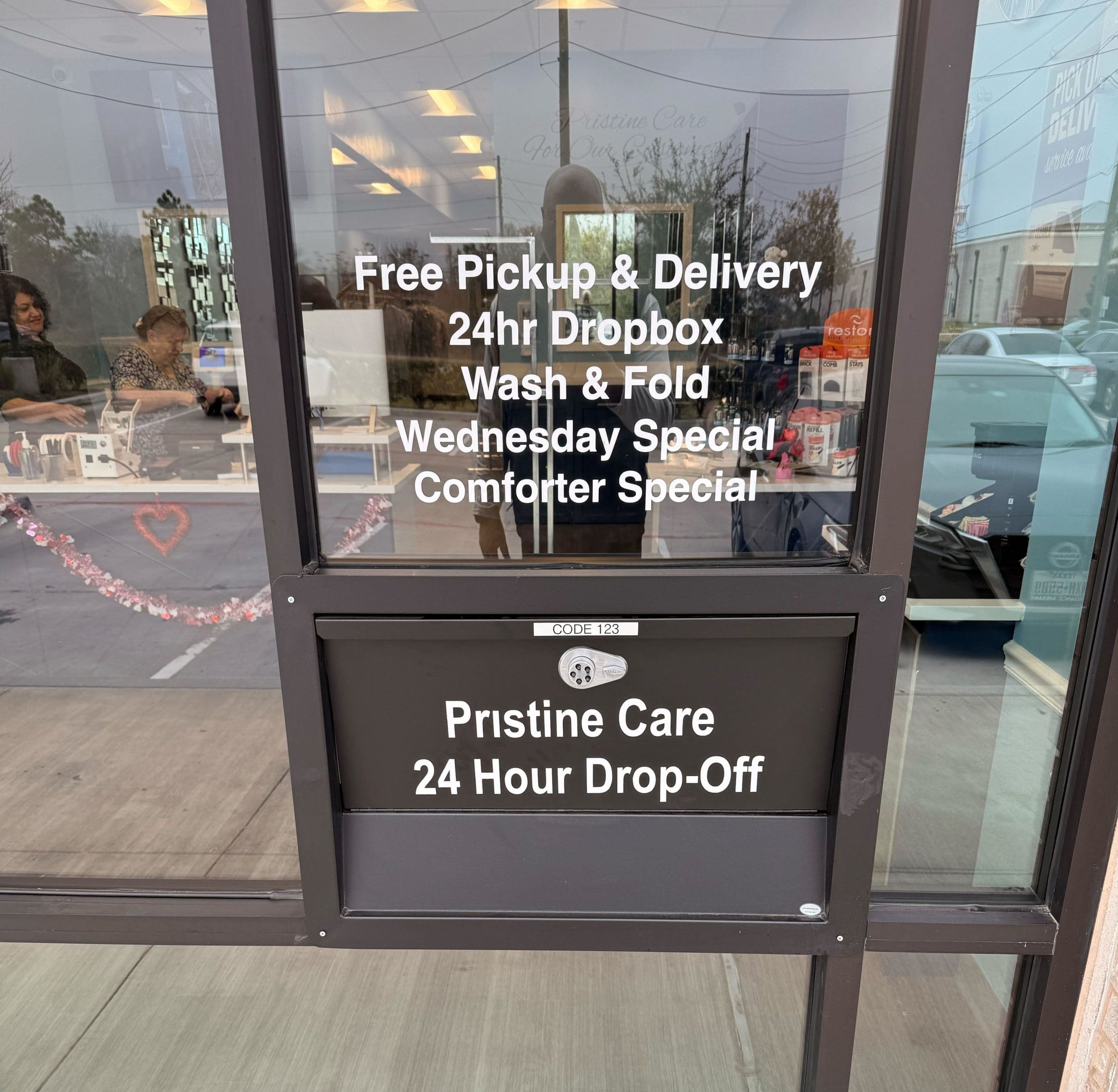 Glass door of a laundromat displaying service details including free pickup, 24hr dropbox, wash and fold, Wednesday and comforter specials, and 24-hour drop-off slot.
