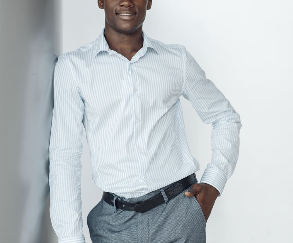 A man wearing a light blue striped dress shirt and gray trousers stands indoors with one hand in his pocket, smiling slightly.