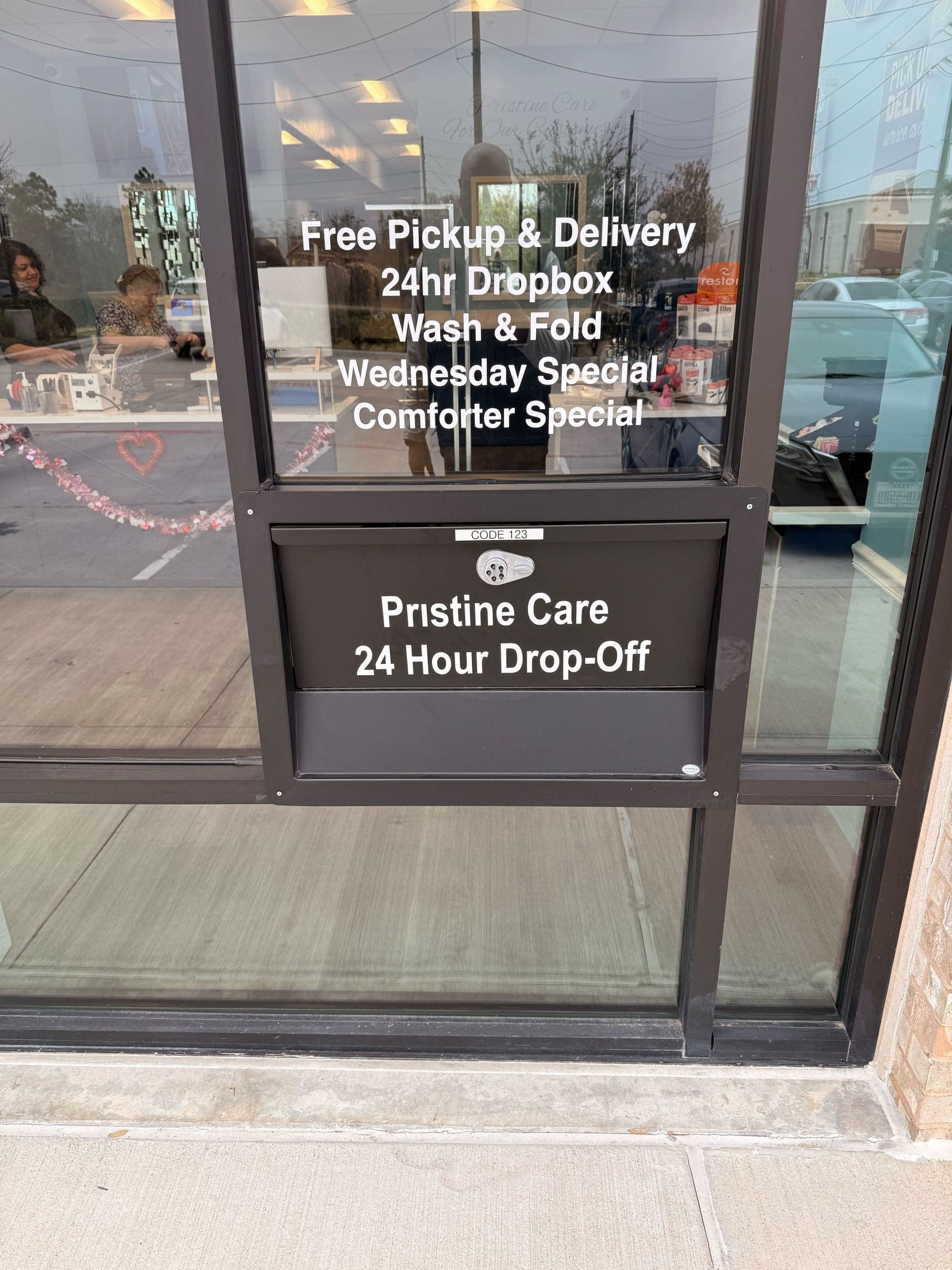 Glass door of a laundromat displaying services such as free pickup and delivery, 24-hour dropbox, wash and fold, and special offers. A drop-off slot is labeled "Pristine Care 24 Hour Drop-Off.