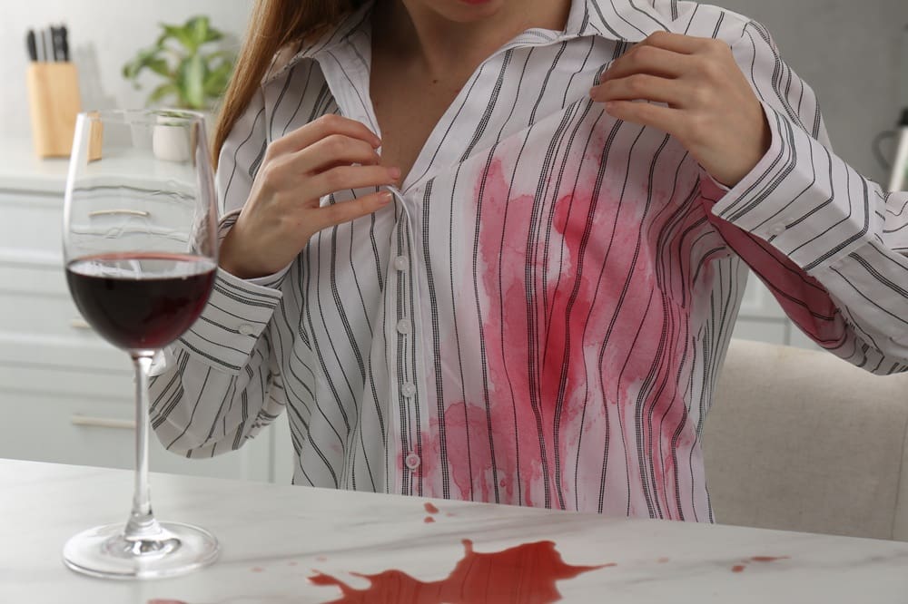 A person wearing a striped shirt examines a red wine stain on their shirt, with a spilled glass of red wine on the table in front of them.